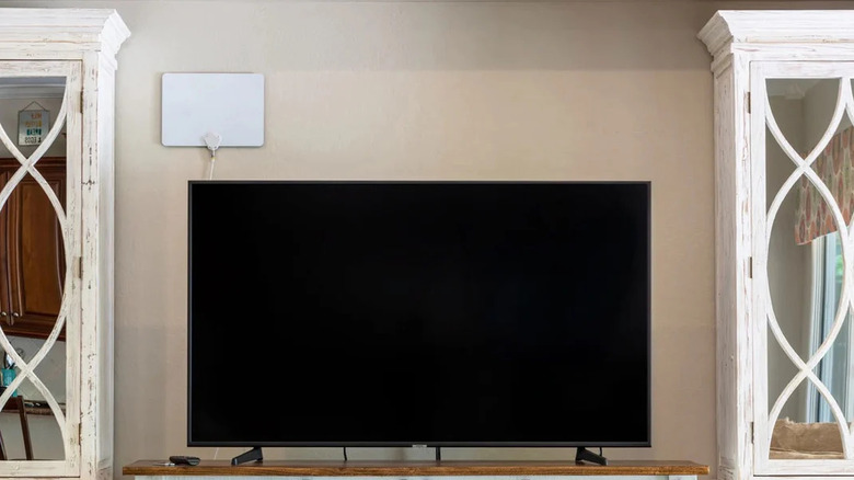 A white indoor antenna placed high on a wall behind a TV placed on a wooden surface