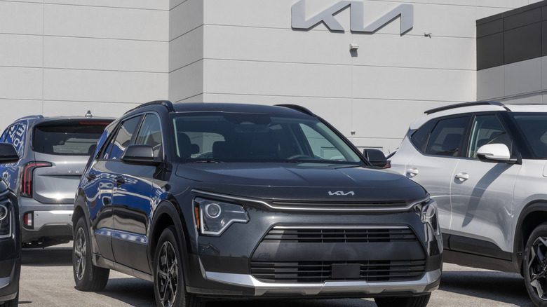 A black Kia Niro Hybrid SUV on display outside a Kia dealership with the Kia logo on a building in the back,