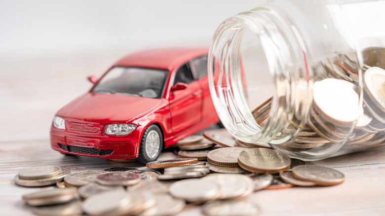 Red model toy car next to a jar filled with coins, spilling onto a wooden surface