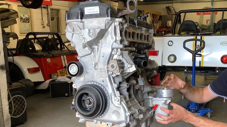 A man's hands are beside a bare Ford Duratec engine on a stand in an auto shop full of classic sports cars.