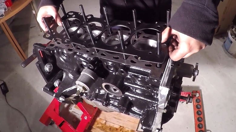 Someone's hands are placing a head gasket onto a bare Mazda BP four-cylinder engine block that's mounted on a stand in a home garage.