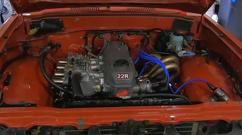 A red Toyota pickup with the hood popped open showing off its detailed 22R engine, while sitting in a home garage workshop.