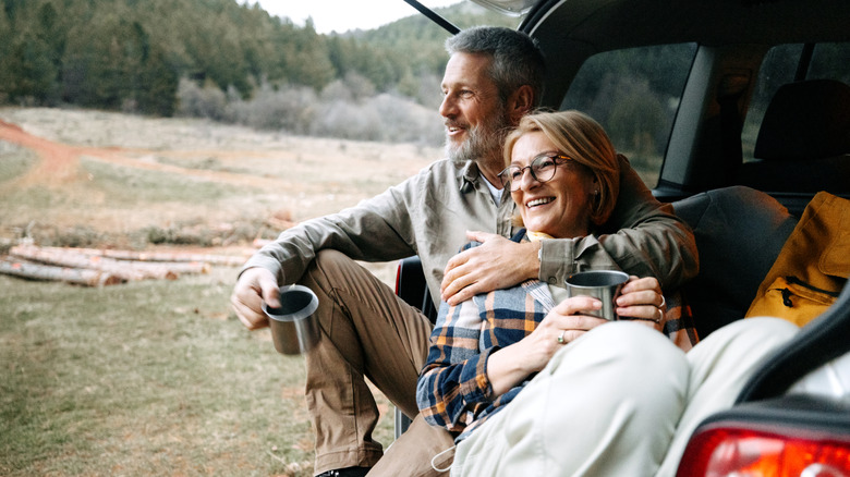 Older couple enjoying cups of coffee while sitting on the back tailgate of an SUV