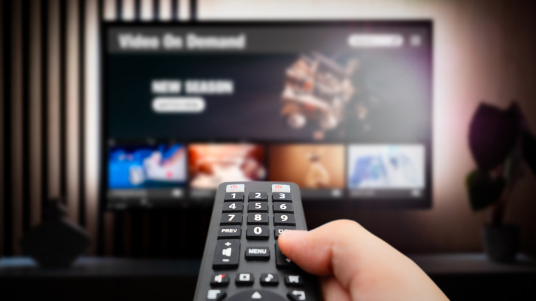 A hand holding a black remote control pointed at a TV displaying a movie streaming platform.