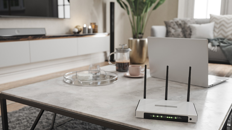A white Wi-Fi router with three antennas on a coffee table next to a laptop with a TV in the background.