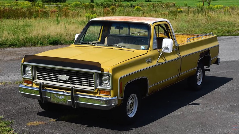 A yellow 1974 Chevrolet C10 Custom 3MT with a two-tone white roof and a noticeably rusty bed sits on a quiet road surrounded by open fields, looking like it just rolled out of a barn find.