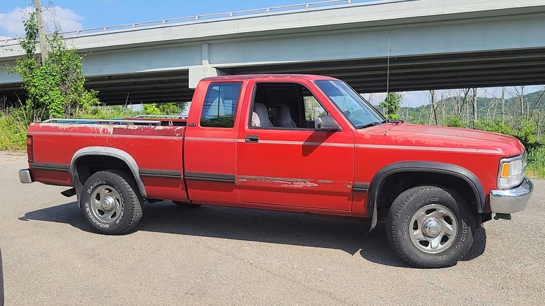 A red Dodge Dakota Club Cab with some honest road wear, faded paint on the hood and some scrapes along the bed parked roadside under a highway overpass on a sunny day.