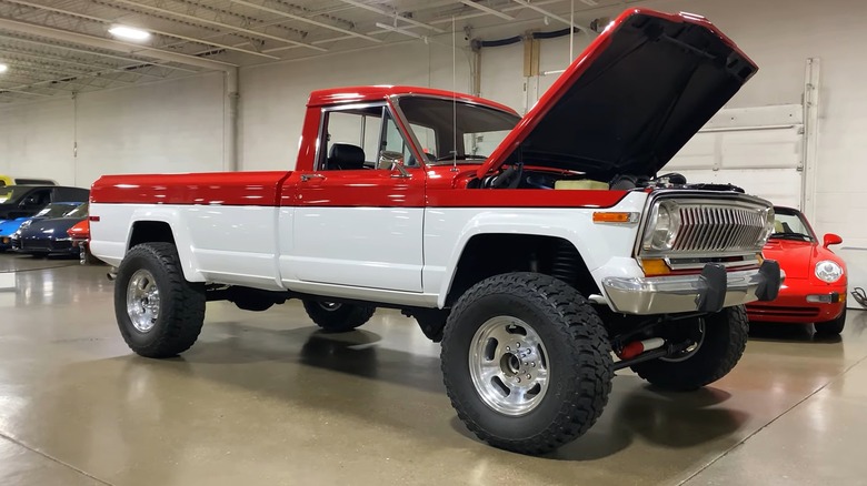 A lifted two-tone red-and-white 1987 Jeep J20 sits inside a dealer showroom with its hood propped open, sitting on massive off-road tires and polished billet wheels.