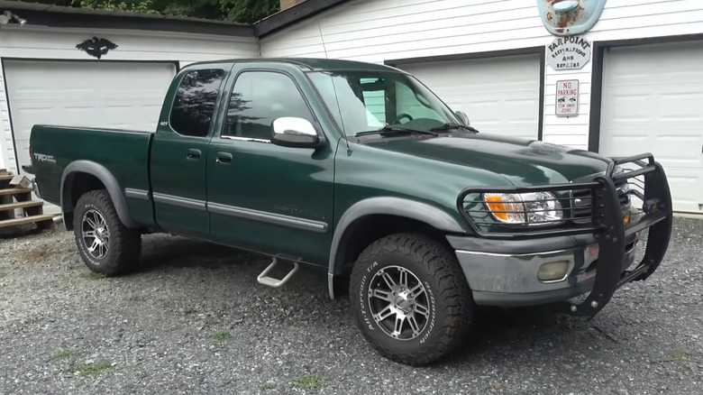 A dark green first-gen Toyota Tundra Access Cab with aftermarket wheels and a brush guard up front sits on a gravel lot outside what looks like a small restoration shop.
