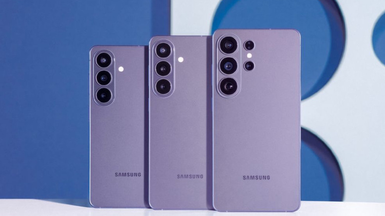 The purple Samsung S26-series phones standing upright without help on a table, with a white and blue wall in the back.