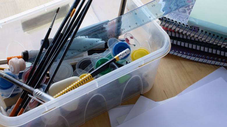 An unorganized bin of art supplies on a wooden desk