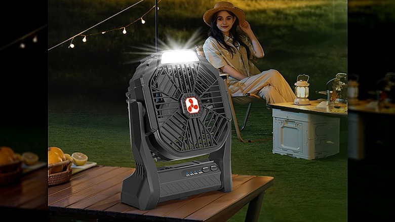 Portable fan placed on a camping table with a woman sitting in the background.
