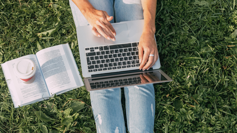 Person sitting on grass holding a laptop with an open book and coffee beside them