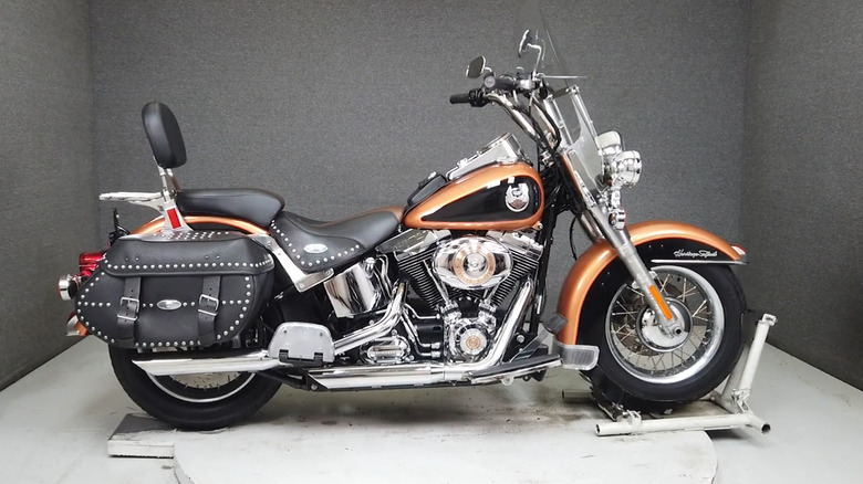 Harley-Davidson Heritage Softail Classic 105th Anniversary Edition motorcycle in Anniversary Copper and Black with commemorative badging, windshield, leather saddlebags, and passenger backrest, displayed indoors