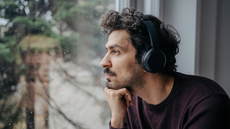 A person with a pensive look gazing out of a window with drops of water on it while wearing an on-ear headphone