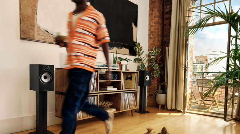 A man walking past a television, with two bookshelf speakers on either side of a packed TV unit in a room attached to a balcony that's open