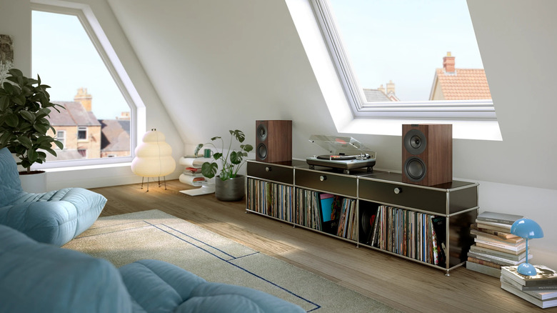 Two KEF Q Concerto Meta 5.1 bookshelf speakers placed on either side of a gramophone on a drawer in the attic, with two blue cushiony seats in front of it