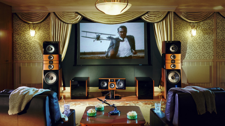 An ornate home theater room, with speakers, snacks on a table, two leather seats on either side, and a projector screen