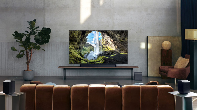 The Samsung HW-Q990F sound system placed in a minimalist living room with a TV, a tall potted plant, and a couch