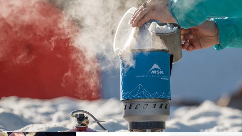 Hands pouring snow into a steaming portable campfire stove