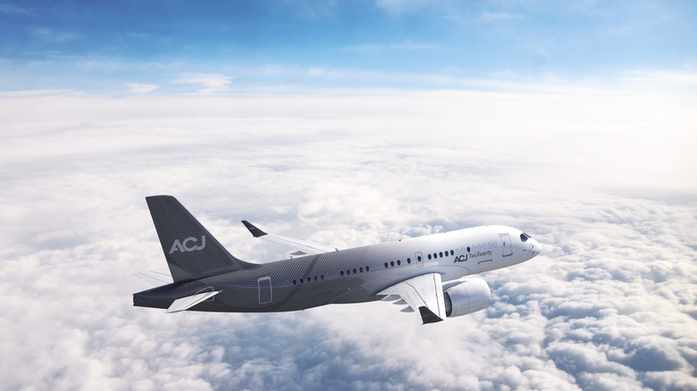A white unliveried Airbus ACJ TwoTwenty private jet cruising above a layer of clouds with a clear blue sky in the background.