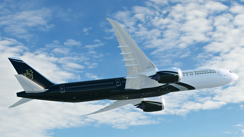 A dark navy and white Boeing BBJ 777X series plane photographed from below while taking off against a blue cloudy sky.