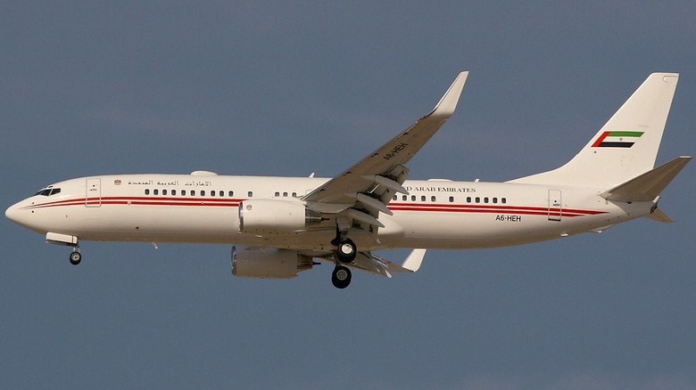A white Boeing BBJ2 used by an operator in Dubai with a red stripe running down the side of the plane, mid-flight.