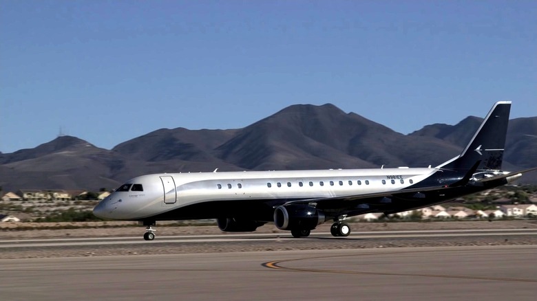 A silver and gray Embraer Lineage 1000E private jet taxiing at a local airport runway, shining in the sun.