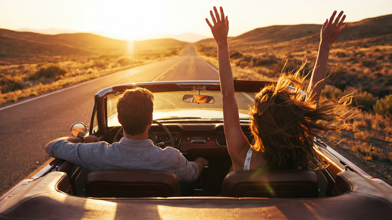 Two people driving a convertible toward the sun.