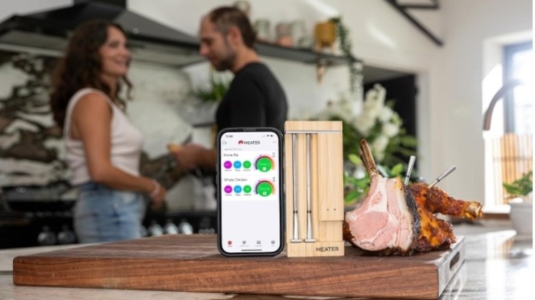 Couple in a kitchen background with an app and meat thermometer in the foreground