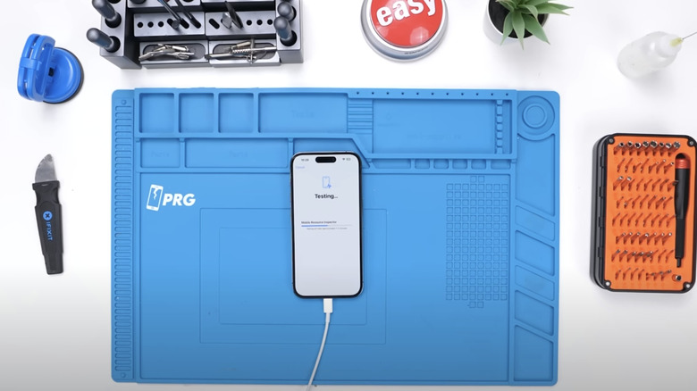 An iPhone 14 Pro Max showing the Testing feature on its display while placed atop a blue workstation for phone repairs, as different tools surround the setup.