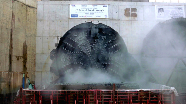 Large cutting wheel breaking through tunnel wall amid cloud of smoke and dust