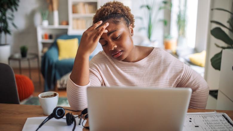 A woman is seated at a home office desk in front of a laptop, her hand on her head in a gesture of concern or stress.