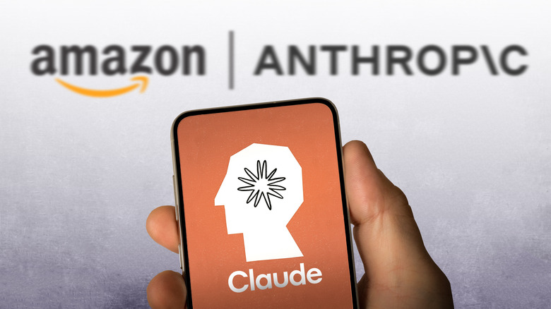 Close up of a hand holding a smartphone showing the Claude logo. In the background, slightly out of focus, are the Amazon and Anthropic logos
