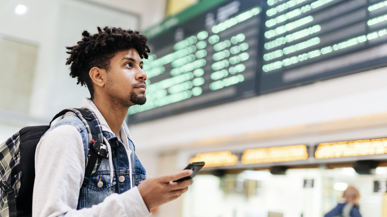 A young man holding a mobile phone looking at the departures of planes in an airport.