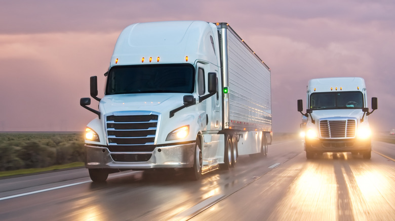 Two semi trucks driving on a wet highway in the evening.