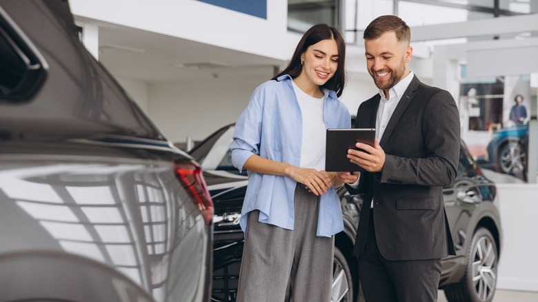 A female customer and male salesperson stand next to a gray car in a cardealership. He is holding a tablet and they are both looking at it and smiling.