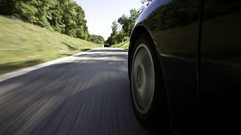 A close-up shot of a car tire in motion on a road, with blurred pavement and trees emphasizing speed and movement.