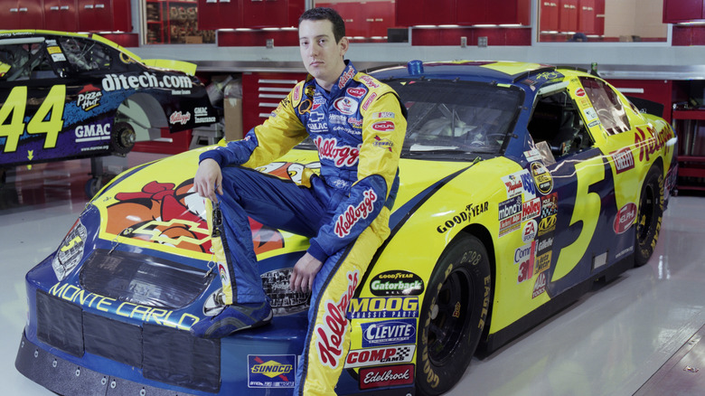 Nascar driver Kyle Busch sitting on hood of blue and gold No. 5 Chevy race car in garage