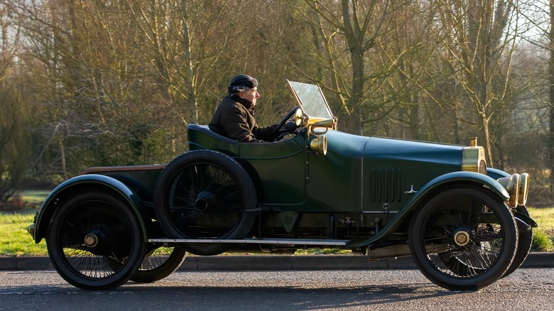 Side shot of man in leather cap driving green Ford Model T