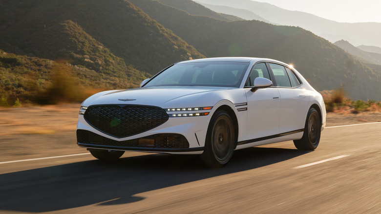 White 2026 Genesis G90 driving down the road, front 3/4 view, mountains in the background