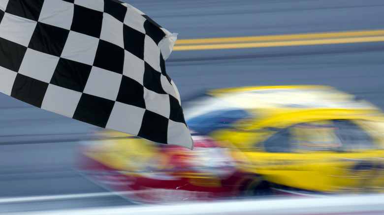 NASCAR stock car racing on-track, blurred, with checkered flag displayed in foreground