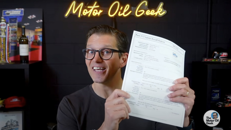 A man in glasses holds up a printed Safety Recall Report in front of a studio backdrop with a sign reading "Motor Oil Geek".