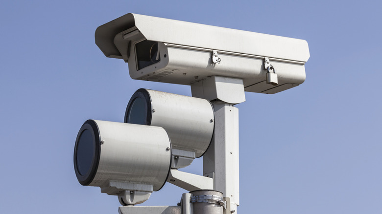 Close-up of a red light traffic camera mounted on a pole against a blue sky.