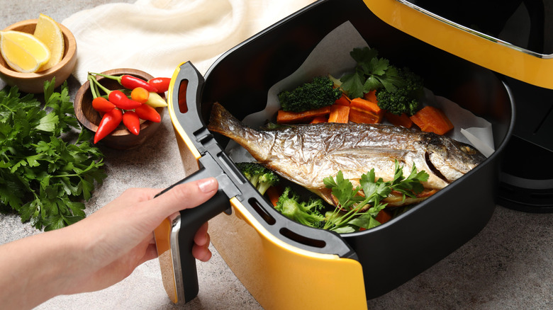 Woman opening air fryer with grilled fish and vegetables at grey table, closeup