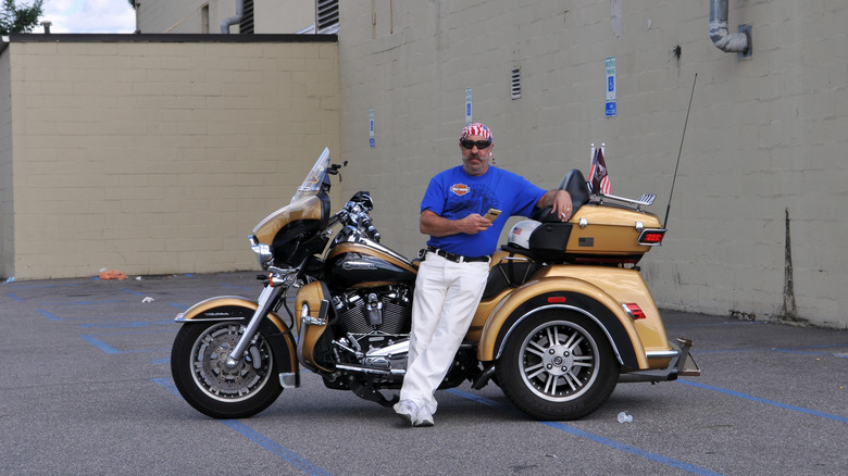 A man leans against his Harley Davidson Trike Tri Glide motorcycle in parking lot
