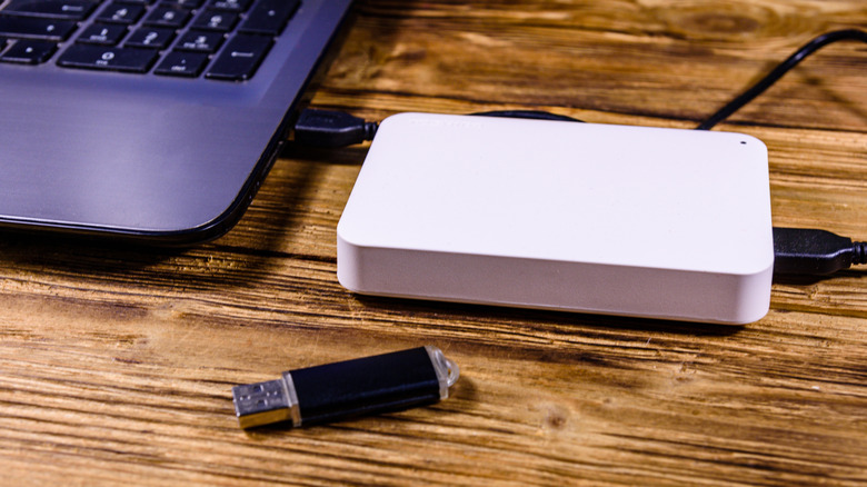 A laptop with an SSD plugged into it and a usb thumb drive sitting next to it, all on top of a wooden desk