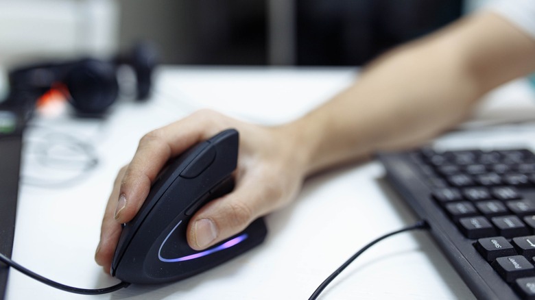 A hand gripping a black vertical mouse on a white desk with PC accessories in the background