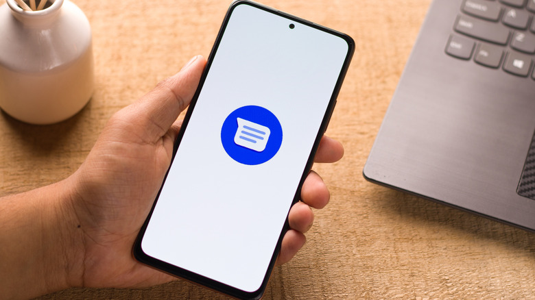A person holding a phone with the Google Messages logo on display; a laptop and small white vase are seen at the back.