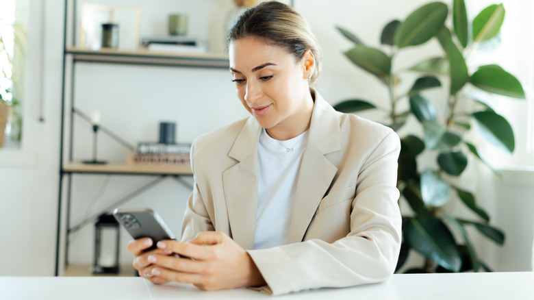 A woman in corporate attire using her phone while sitting on a desk with plants and a shelf of different items are seen at the back.